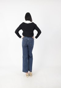 Frau mit langen dunklen Haaren steht mit dem Rücken zur Kamera, trägt einen schwarzen Pullover mit weißem Rüschenkragen, blaue Jeans und cremefarbene High-Heels.