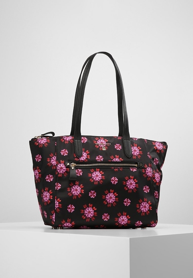 Sac cabas noir avec un motif floral rose et rouge, deux longues poignées noires, et une poche zippée à l'avant sur une surface blanche.