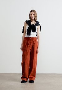 Femme debout portant un débardeur blanc, un pantalon large couleur rouille avec des poches, un pull noir posé sur les épaules et des chaussures noires.