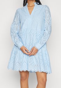 Ljusblå klänning gjord av broderad bomull, med långa ärmar, V-ringning och en lager-på-lager kjol med vågformad nederkant och intrikata utskärningar.