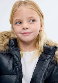 Černá péřová bunda s kapucí lemovanou kožešinou, s zipovým zapínáním a prošívanými sekcemi, nošena přes bílou košili. Světle blond vlasy.