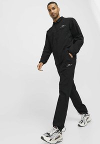 Melna zip-up jakna ar sānu kabatām, kopā ar melnajām biksēm, kurām ir logo akcenti. Modelis valkā retro stila sporta apavus ar dažādām krāsām.