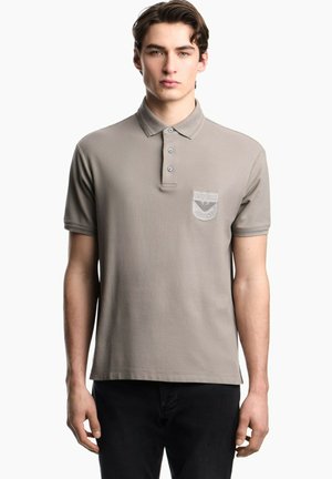 Jonge man die een taupekleurig poloshirt met korte mouwen en een borstzakembleem draagt en zwarte broek, die vooruit kijkt tegen een witte achtergrond.