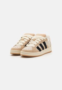 Baskets en daim beige avec quatre rayures noires, lacets crème et tige texturée. Présente des perforations sur la pointe et une semelle en caoutchouc.