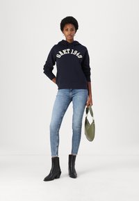 Sweat à capuche bleu marine avec l'imprimé "GANT 1949", jean slim bleu, bottines noires, et un sac à main vert. Matière lisse, coupe décontractée.