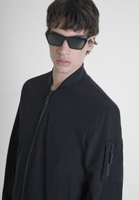 Chaqueta bomber negra con cremallera al frente y cuello acanalado. Cuenta con un bolsillo con cremallera en la manga izquierda. Textura de tela suave, diseño minimalista.