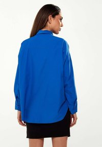 Blue button-up shirt with long sleeves, collar, and rounded hem. Made of smooth fabric; features cuff buttons and a relaxed fit.