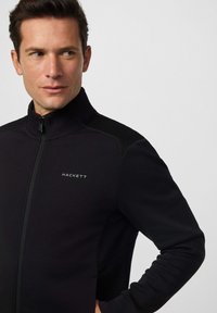 Muškarac u crnoj jakni na zip s logom "HACKETT" na prsima, gleda lijevo, nasuprot jednostavnoj svjetlosivoj pozadini.