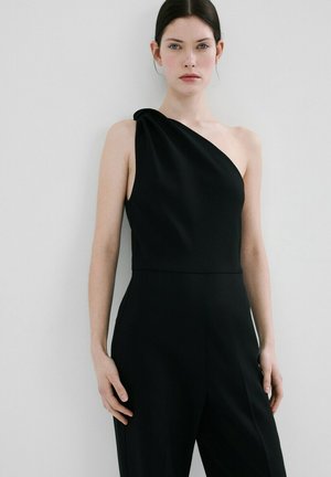 Femme portant une combinaison noire sans manches avec un décolleté asymétrique à une épaule, debout devant un fond blanc uni.