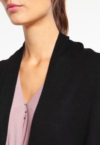 Svart stickad cardigan med öppen front, mjuk textur och medel tjocklek. Bärs över en ljusrosa topp med knappar vid halsringningen.