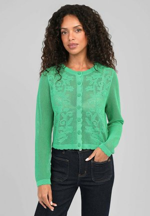 Femme aux cheveux bouclés portant un cardigan vert à motifs boutonné et un jean foncé, debout devant un fond gris uni.