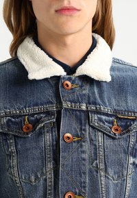 Jeansjacka med vit faux shearling-krage, kopparfärgade knappar och två bröstfickor. Har en klassisk blåtvätt och synliga sömnadsdetaljer.