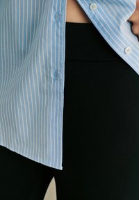 Világoskék csíkos ing gombos záródással, fekete leggings-szel párosítva. Az ingnek finom fényessége van és ívelt hemje.