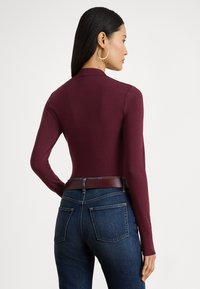 Hosszú sötét hajú nő, nagy karika fülbevalóval, testhezálló bordó hosszú ujjú felsőben, sötétkék farmerben és hozzá illő övvel, oldalra fordulva.