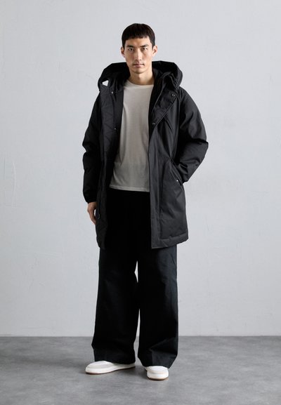 Schwarze isolierte Parkajacke mit Kapuze, die über eine gesteppten Struktur, vordere Druckknöpfe und zwei seitliche Taschen verfügt. Kombiniert mit weiten schwarzen Hosen.