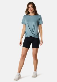 Ljusblå kortärmad topp med knuten frontdesign, matchad med svarta cykelshorts. Tyget verkar slätt och sträckt. Vita sneakers.