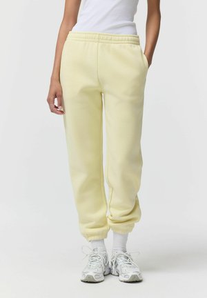 Kobieta ubrana w jasnożółte spodnie dresowe z elastycznym pasem i ściągaczami, białe skarpetki oraz białe buty sportowe, stojąca na jednolitym tle.
