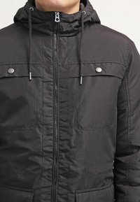 Svart huva jacka med zip-fram, två bröstfickor med klaffar och dragsnoddar vid huvan. Slät textur, vadderad design.