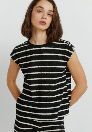 Kobieta ubrana w czarno-biały, bezrękawnik w paski z guzikami na jednym ramieniu, w zestawie z pasującymi spodniami w paski, stojąca z rękami za plecami.