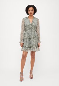 Short green dress with a floral pattern, layered ruffles, V-neck, and sheer long sleeves. Worn with silver strappy heels.
