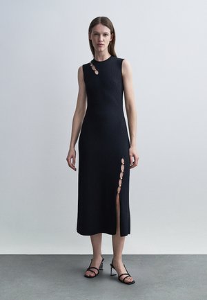 Femme debout portant une robe midi noire sans manches en maille côtelée avec des accents d'anneaux dorés sur l'épaule et une fente sur la cuisse, chaussée de sandales noires à talons.