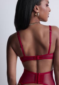 Ensemble de lingerie rouge présentant un design transparent avec des bretelles réglables et une fermeture à crochets, mettant en valeur un tissu lisse et une forme structurée.