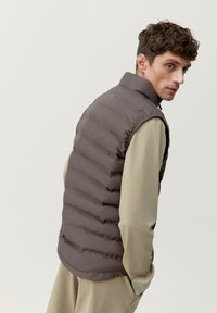 Gevoerde vest in donkerbruin met gewatteerde details, een hoge kraag en een gladde textuur. Draag dit over een lichtbeige top met een minimalistisch ontwerp.