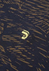 Gebreid stof in marineblauw met een draaiend bruine patroon. Bevat een klein, gele geborduurd logo met een gebogen ontwerp.