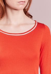 Haut rouge à manches longues avec un col rond, présentant une bordure blanche contrastante le long du bord et une texture lisse côtelée.