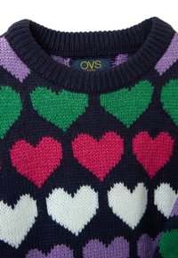 Strickpullover mit navy Hintergrund, der farbenfrohe Herzmuster in Grün, Pink, Weiß und Lila zeigt. Rippstrickdetail am Ausschnitt sichtbar.