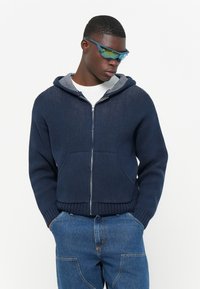 Marineblå hettegenser med glidelås, ribbestrikkede mansjetter og hem, teksturert kropp, frontlommer og hette med hvit fôr, sammen med denimbukser.