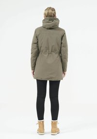 Olivgrön parkajacka med dragsko i midjan, huva och elastiska ärmslut. Bärs över svarta leggings och tanfärgade ankelstövlar. Enkelt design.