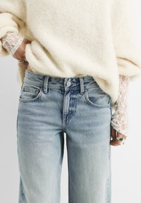 Jeans in denim blu chiaro a vita alta, con tasche laterali, un bottone frontale e una leggera scoloritura. Indossati con un pullover crema e dettagli in pizzo.