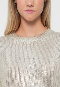 Jasnosrebrny sweter dzianinowy z okrągłym, prążkowanym dekoltem, charakteryzujący się metalicznym połyskiem i teksturowanym wyglądem. Brak widocznych wzorów lub ozdób.