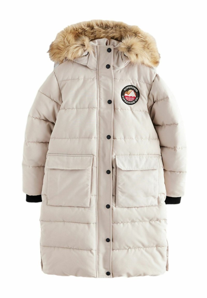 Helle beige Steppjacke mit einer Kapuze aus Kunstfell, schwarzen Knöpfen, zwei Eingrifftaschen und einem runden Logo-Patch auf der Brust. Mit gerippten Bündchen.