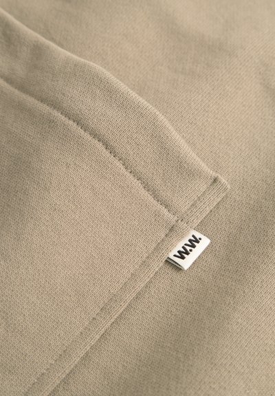 Beige szövet lágy, texturált felülettel és látható varrással, a szegélyén egy kis fehér címkével, fekete felirattal.