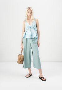 Haut peplum bleu clair avec un ourlet volanté et des bretelles, associé à un pantalon palazzo vert menthe ample. Le mannequin tient un sac en osier. Sandales noires.
