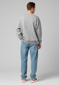 Felpa grigia chiara con spalle montate, abbinata a jeans azzurri chiari con dettagli sulle tasche posteriori. Il modello indossa sneaker bianche.