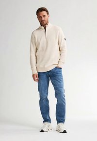 Cremefarbener, strukturierter Pullover mit Reißverschluss am Kragen, kombiniert mit hellblauen Jeans und weißen Sneakers. Mit einem Logo auf dem Ärmel akzentuiert.