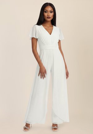 Frau trägt einen weißen Overall mit weitem Bein, Flatterärmeln und V-Ausschnitt, posiert vor einem beigen Hintergrund mit High-Heel-Sandalen.