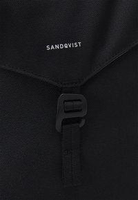 Schwarze Tasche aus strapazierfähigem Stoff mit einem Klappenverschluss und einer schwarzen Schnalle. Das "SANDQVIST"-Logo ist in Weiß auf der Vorderseite aufgedruckt.