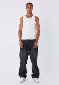 Canotta bianca con logo nero, jeans larghi scuri, catena d'argento e sneakers bianche. Modello in piedi dritto su uno sfondo neutro.
