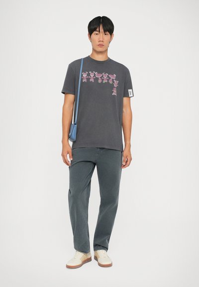 Maglietta grigia con una grafica di figure rosa in stile cartone animato, abbinata a jeans grigi scuri e scarpe chiare. Inclusa borsa a tracolla blu.
