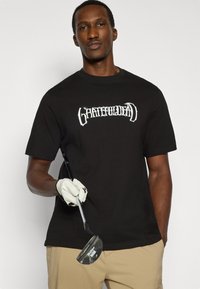 Μαύρο βαμβακερό t-shirt με λευκό στυλιζαρισμένο λογότυπο κειμένου "Grateful Dead" στο μπροστινό μέρος. Το μοντέλο κρατά ένα γκολφ ρόπαλο και φοράει λευκό γάντι.