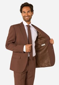 Homme en costume marron et cravate souriant, tenant sa veste ouverte pour révéler la doublure intérieure et l'étiquette de la marque.