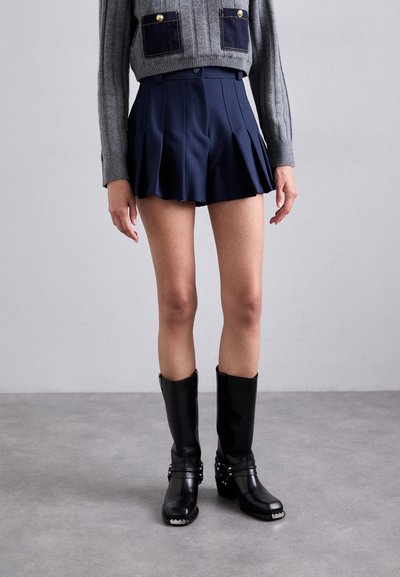 Marineblaue, plissierte Shorts aus weichem Stoff, mit hoher Taille und A-Linien-Form. Kombiniert mit kniehöhen schwarzen Lederstiefeln mit metallischen Akzenten.