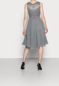 Robe grise avec un corsage en dentelle, sans manches, et une jupe fluide longueur genou. Portée avec des chaussures noires à talons hauts. Vue de dos.