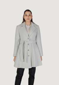 Cappotto grigio testurizzato con vita arricciata, reverse affilati, chiusura con bottoni e due tasche frontali. Abbinato a pantaloni scuri.