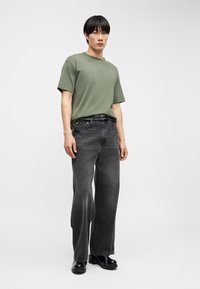 Νεαρός άνδρας όρθιος, φοράει πράσινο κοντομάνικο πουκάμισο, ξεθωριασμένο μαύρο τζιν με φαρδιά πόδια, μαύρα παπούτσια με τακούνι και μαύρη ζώνη με καρφιά, πάνω σε απλό φόντο.