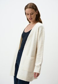 Cardigan lungo e aperto in tessuto texturizzato color crema. Presenta due tasche frontali e polsini e orlo a costine.
